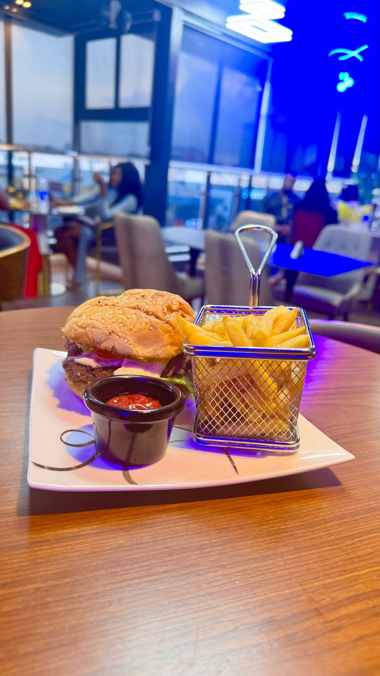 A gourmet burger meal with fries in a wire basket and dipping sauce served on a white plate in a modern restaurant with blue ambient lighting