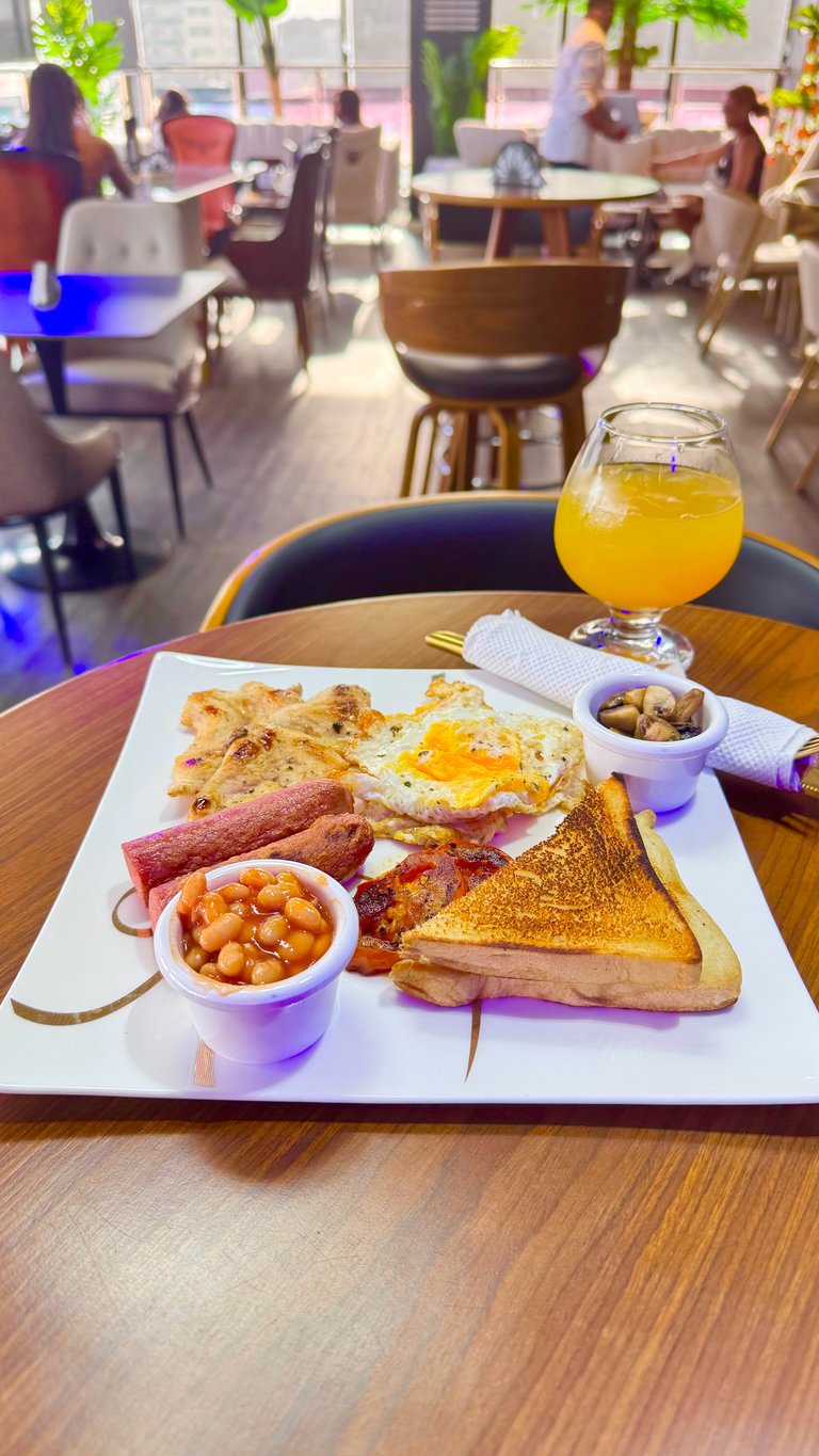 Full English breakfast plate with eggs, bacon, sausage, toast, and baked beans, served at a bright café with orange juice