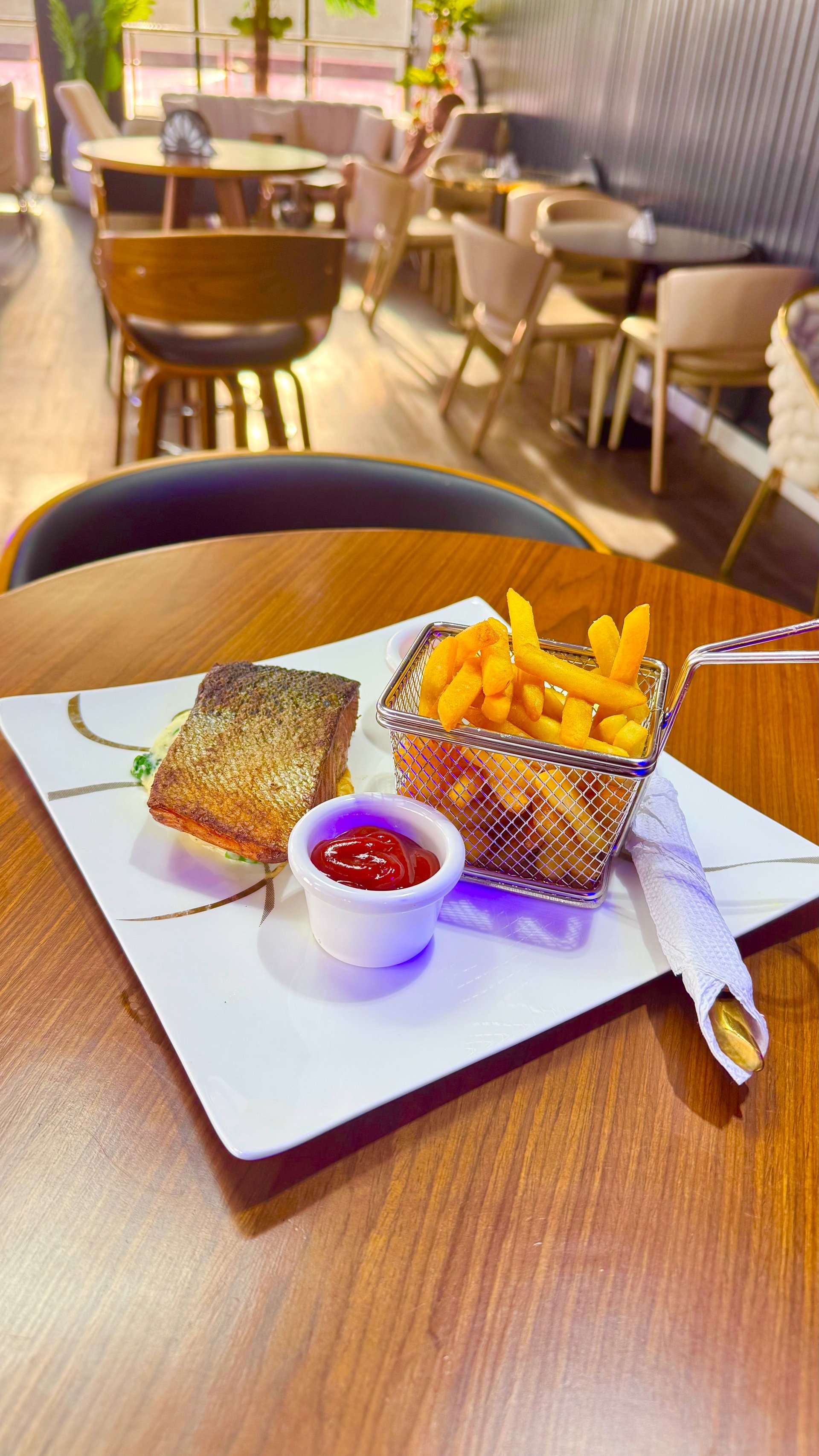 Plated meal with crispy fish fillet, golden fries in a metal basket, and ketchup in a white cup on a wooden table in a modern restaurant